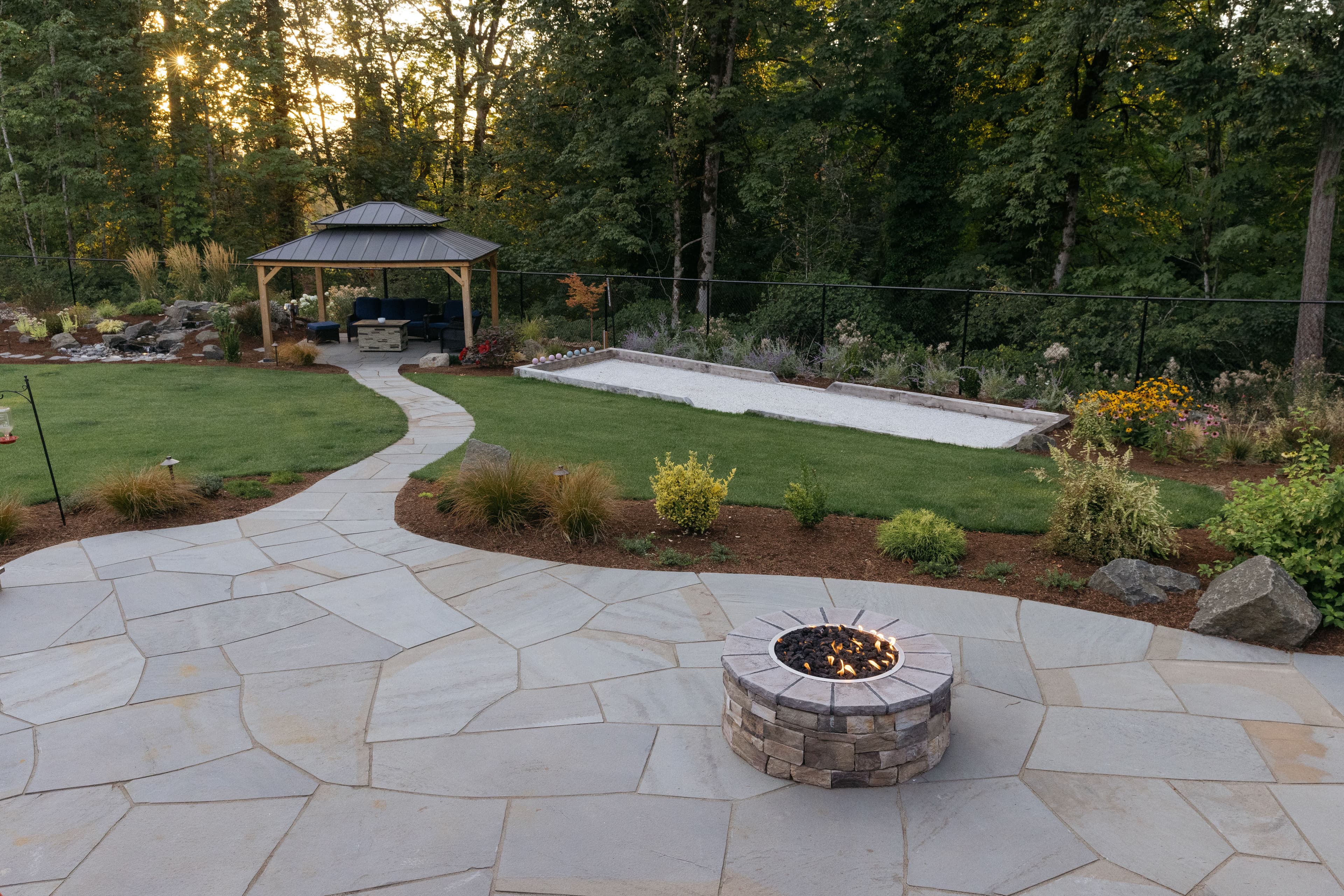 Backyard landscape with a fire pit, gazebo, and landscaped garden area at sunset.